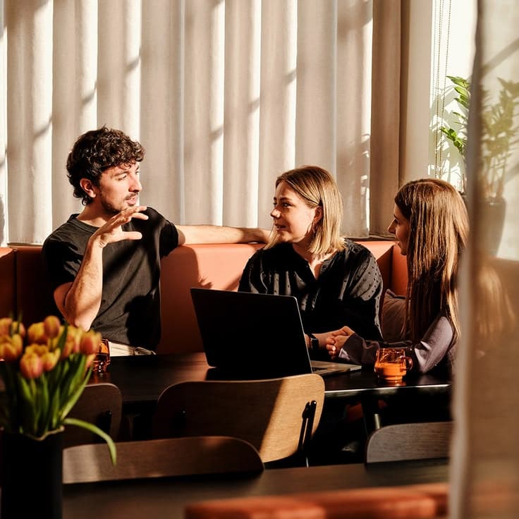 Three people in conversation at a table, warm afternoon light through sheer curtains, laptop open between them